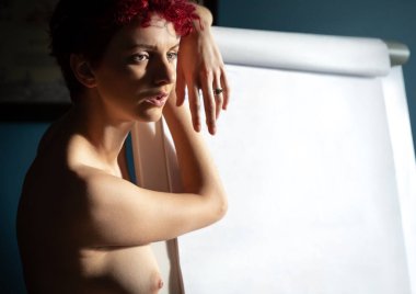 Kısa kızıl saçlı çıplak bir kadın kapalı bir ortamda boş bir tuvale yaslanıyor..