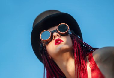 Göz kamaştırıcı bir kız kırmızı mayo, siyah şapka ve parlak mavi gökyüzünün altında kaynak gözlüğü giyerken kızıl rastalarını sergiliyor. Sahne yaratıcı metin için boş alan sunuyor.