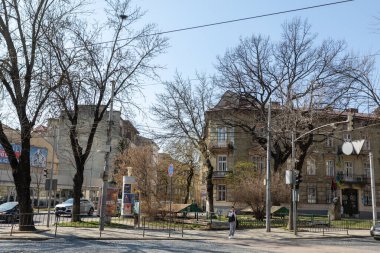 Lviv, Ukrayna - Mar. 22, 2025: Ukrayna 'nın Lviv şehrinde yayaların tarihi binaların ve çıplak ağaçların yanından geçtiği canlı bir bahar sabahı. Güneş sahnenin üzerine ılık bir parıltı saçar..