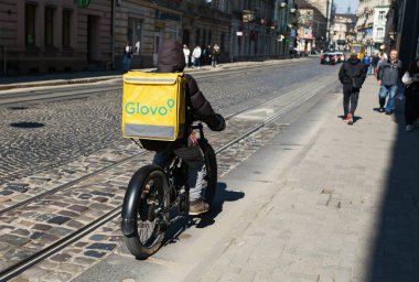 Lviv, Ukrayna - Mar. 22, 2025: Bir bisikletçi Lviv, Ukrayna 'nın canlı sokaklarında şehir yaşamını gözler önüne sererek yiyecek dağıtıyor. Çakıl taşları ve yayalar canlı bir atmosfer yaratıyor..