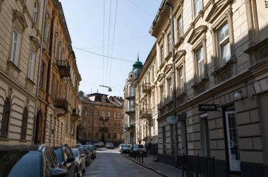 Lviv, Ukrayna - Mar. 22, 2025: Ukrayna 'nın Lviv şehrindeki tarihi binalarla çevrili büyüleyici kaldırım taşı caddesi. Parlak güneş ışığı, paralel park etmiş arabalar modern bir dokunuş eklerken, karmaşık cepheleri vurgular..