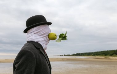 Yüzü kumaşla kaplı, şapka ve ceket giyen bir adam, deniz kıyısında ellerini kullanmadan ağzında bir elma tutuyor..