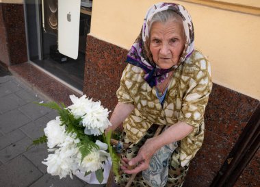 LVIV, UKRAINE - Haziran. 20, 2021: Yaşlı bir kadın sokakta oturuyor, elinde bir buket beyaz çiçekle yoldan geçenlere satıyor. Geleneksel kıyafetleri yerel kültürü ve tarihi yansıtıyor..