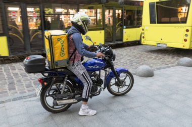 LVIV, UKRAINE - Haziran. 20, 2021: kask takan bir yemek dağıtıcısı, izole edilmiş bir çantayı motosikletle taşıyor. Şehir merkezinde kaldırım taşı sokaklar ve yakındaki sarı bir otobüs yer alıyor..