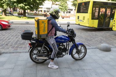 LVIV, UKRAINE - Haziran. 20, 2021: Mavi motosikletli bir yiyecek teslimatçısı sokakta sarı bir sırt çantasıyla yiyecek teslimatına hazırlanırken görüldü. Şehir yayalar ve otobüslerle dolu..
