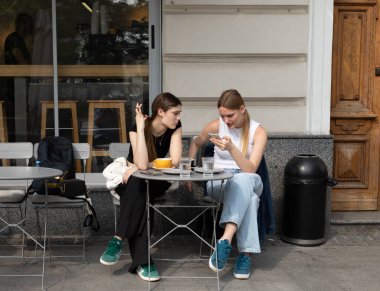 Lviv, Ukrayna - Haziran. Güneşli bir günde iki genç kadın bir kafede oturuyor, biri telefonuna bakıyor..