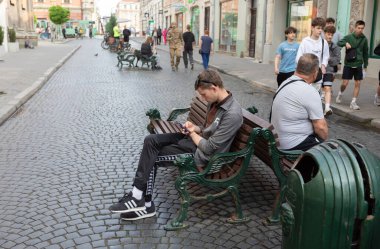 Lviv, Ukrayna - Haziran. 01, 2025: Bir adam bir bankta oturuyor, bir kaldırım taşı sokağında telefonuna dalmış..