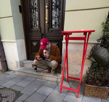 Lviv, Ukrayna - Haziran. 01, 2025: Şapkalı bir adam kırmızı masanın yanında telefonuna bakarken içecekten zevk alır.