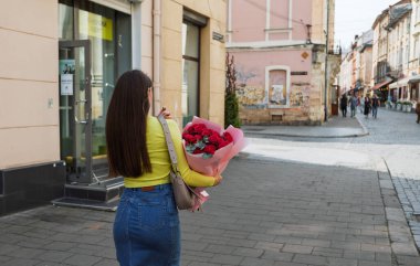 Lviv, Ukrayna - Haziran. 01, 2025: Sarı bluzlu ve kot etekli bir kadın şehir caddesinde bir buket kırmızı gül tutuyor.