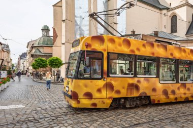 Lviv, Ukrayna - Haziran. 01, 2025: Peynir desenli eşsiz sarı bir tramvay, arka planda insanlar ve tarihi binalar olan bir kaldırım taşı caddesinde ilerliyor..
