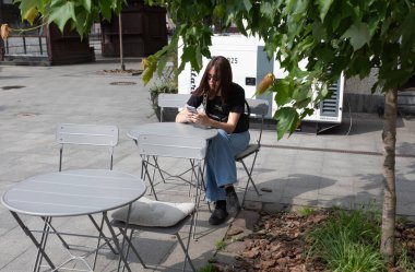 Lviv, Ukrayna - Haziran. 01, 2025: Güneşli bir günde bir kadın kafeterya masasında oturuyor, telefonuna dalmış..