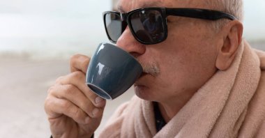 Güneş gözlüğü takan yaşlı bir adam bardaktan yudumluyor, içkisinin tadını çıkarıyor..