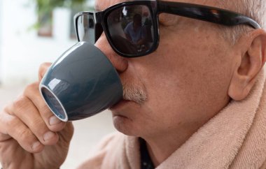 Güneş gözlüklü yaşlı bir adam mavi bardaktan içiyor, rahat ve rahat, açık renkli bir bornozla..