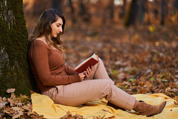 Young woman reading a book outdoor in the forest in the late autumn at sunset