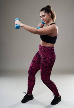 Çekici ve kilolu kadın halterlerle çalışıyor, gri arka planda izole edilmiş.