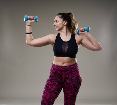 Çekici ve kilolu kadın halterlerle çalışıyor, gri arka planda izole edilmiş.