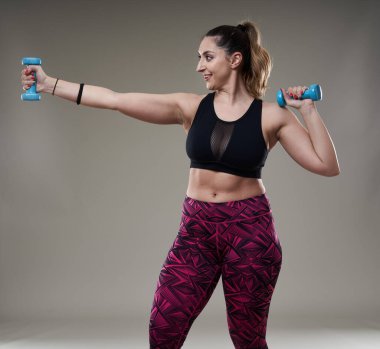 Çekici ve kilolu kadın halterlerle çalışıyor, gri arka planda izole edilmiş.