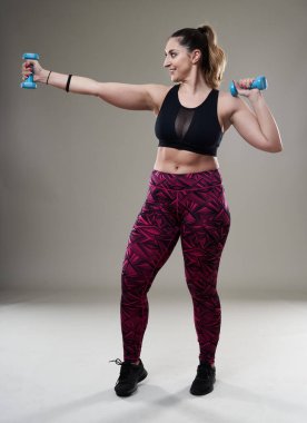 Çekici ve kilolu kadın halterlerle çalışıyor, gri arka planda izole edilmiş.