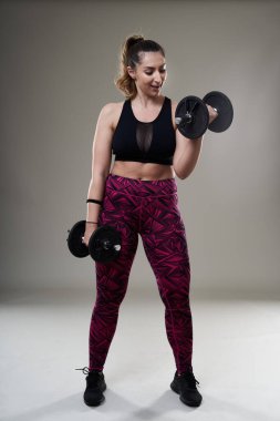Çekici ve kilolu kadın halterlerle çalışıyor, gri arka planda izole edilmiş.