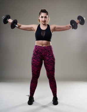 Çekici ve kilolu kadın halterlerle çalışıyor, gri arka planda izole edilmiş.