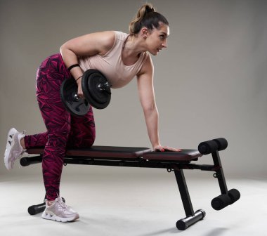 Kendine güveni tam, kilolu bir kadın spor salonundaki halterlerle çalışıyor. Gri arka planda izole edilmiş.