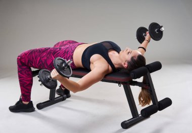 Kendine güveni tam, kilolu bir kadın spor salonundaki halterlerle çalışıyor. Gri arka planda izole edilmiş.