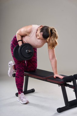 Kendine güveni tam, kilolu bir kadın spor salonundaki halterlerle çalışıyor. Gri arka planda izole edilmiş.