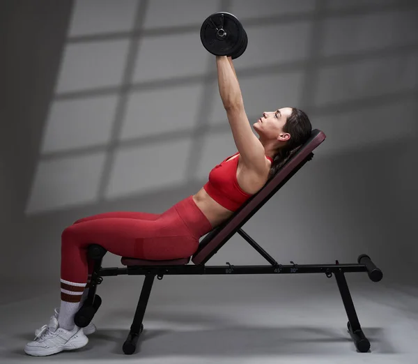 運動能力若いですフィットネスモデル女性で赤いジムの服を着て胸のトレーニングとともにダンベルオン傾斜ベンチ — ストック写真