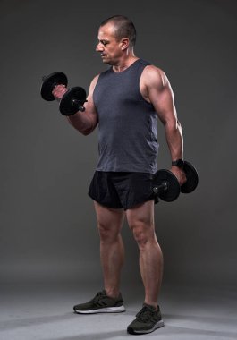 Tişörtlü ve şortlu, gri arka planda spor yapan olgun, beyaz kaslı bir adam.