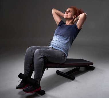 Kafkasyalı olgun bir kadın gri arka plandaki eğimli bankta karın kasları yapıyor. 