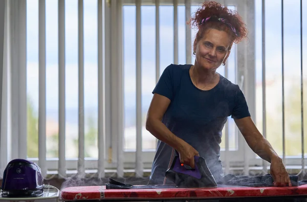 卷发红头发的家庭主妇在窗边熨烫衣服 — 图库照片