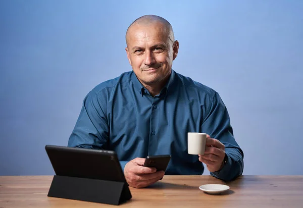 机の前にタブレットを持ってる幸せなビジネスマンコーヒーを飲んでる青い背景の上に — ストック写真