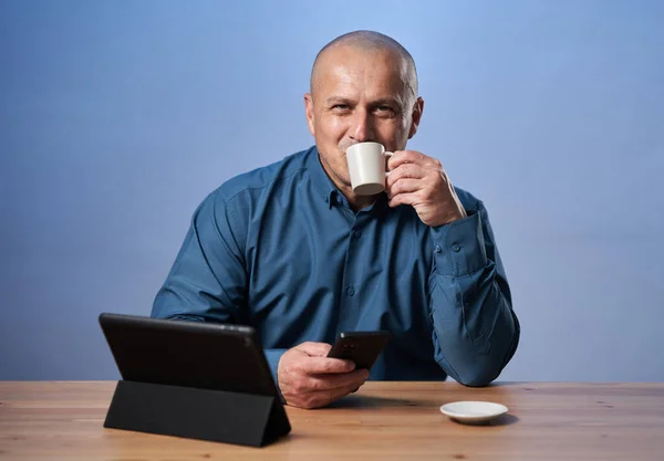 Happy business man drinking coffee having a tablet in front of him at his desk, over blue background