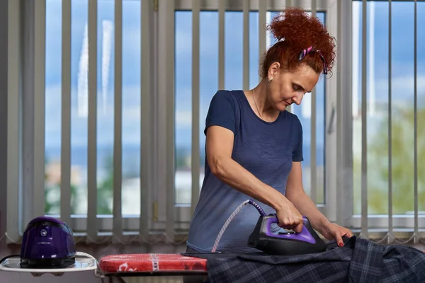 Kıvırcık Saçlı Kızıl Saçlı Hanımı Pencerenin Kenarındaki Kıyafetleri Ütülüyor — Stok fotoğraf