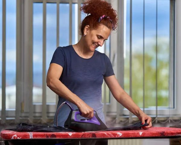 Kıvırcık Saçlı Kızıl Saçlı Hanımı Pencerenin Kenarındaki Kıyafetleri Ütülüyor — Stok fotoğraf
