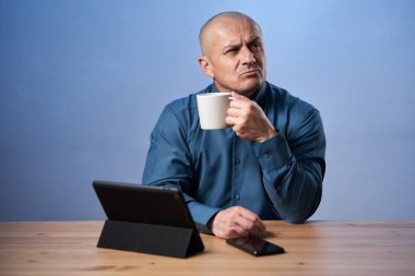 Düşünceli, olgun iş adamı masasında, tabletinin önünde kahve içiyor.