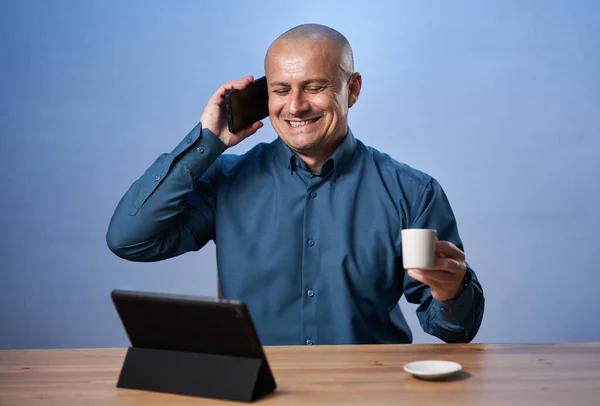 タブレットの前の机にコーヒーを飲みながら携帯電話で話してるビジネスマンだ — ストック写真