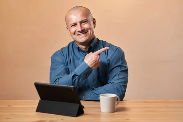 机の上にタブレットとコーヒーカップを持つ幸せな陽気なビジネスマンです 画像の側面に向かって示す 流行の背景の上に — ストック写真