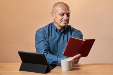 Yetişkin bir iş adamı, işyerindeki masasında kahve molası verip kitap okuyor.