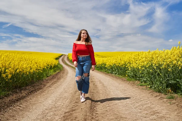 若いですブロンドCaucasian女性で赤トップとジーンズ歩くと未舗装の道を通って咲く菜の花畑で晴れた日 — ストック写真