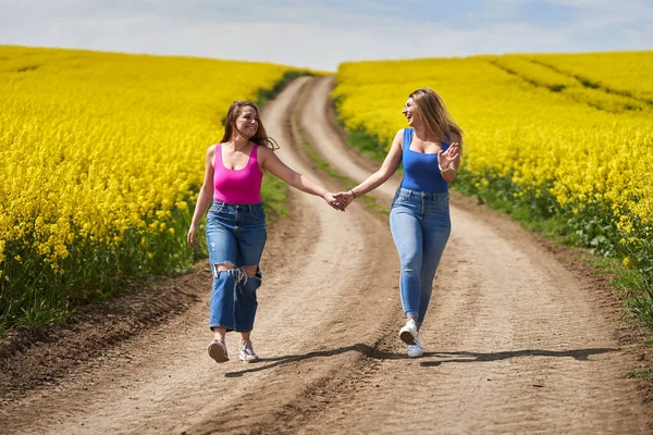 2プラスサイズのガールフレンド喜んで手を取り合って 開花菜の花畑を通って未舗装の道を歩く — ストック写真