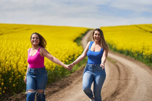 两个大一点的女朋友手牵着手 高高兴兴地走在一条泥泞的路上 穿过一片盛开的油菜地 — 图库照片