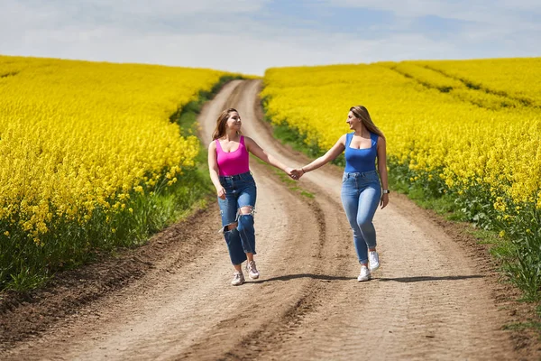 2プラスサイズのガールフレンド喜んで手を取り合って 開花菜の花畑を通って未舗装の道を歩く — ストック写真
