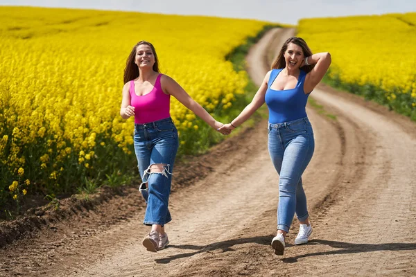两个大一点的女朋友手牵着手 高高兴兴地走在一条泥泞的路上 穿过一片盛开的油菜地 — 图库照片