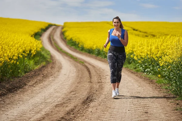プラスサイズ美しいラテン女性ジョギング未舗装の道路でカノーラフィールド — ストック写真