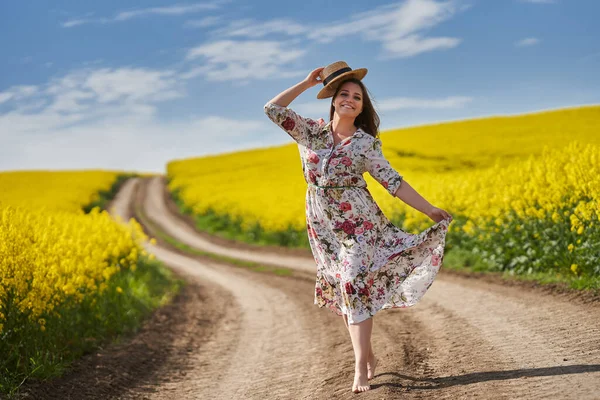 花のドレスを着たプラスサイズの豪華な女性は 田舎の花の菜の花畑によって未舗装の道路を裸足で走り — ストック写真