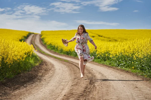 花のドレスを着たプラスサイズの豪華な女性は 田舎の花の菜の花畑によって未舗装の道路を裸足で走り — ストック写真
