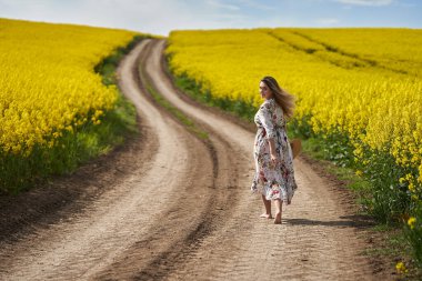 Çiçekli elbiseli, sarışın, romantik bir kadın. Kanola tarlasında çıplak ayakla yürüyor.