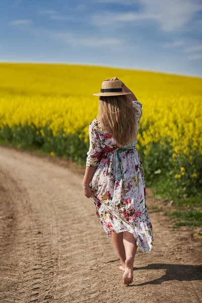 穿着花衣 赤着脚在一条穿过油菜田的土路上行走的浪漫而美丽的金发碧眼的慌张的年轻女子 — 图库照片