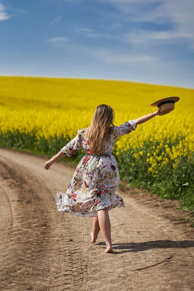 穿着花衣 赤着脚在一条穿过油菜田的土路上行走的浪漫而美丽的金发碧眼的慌张的年轻女子 — 图库照片